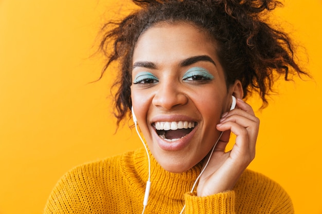 Enthousiaste femme africaine portant un chandail debout isolé, écoutant de la musique avec des écouteurs