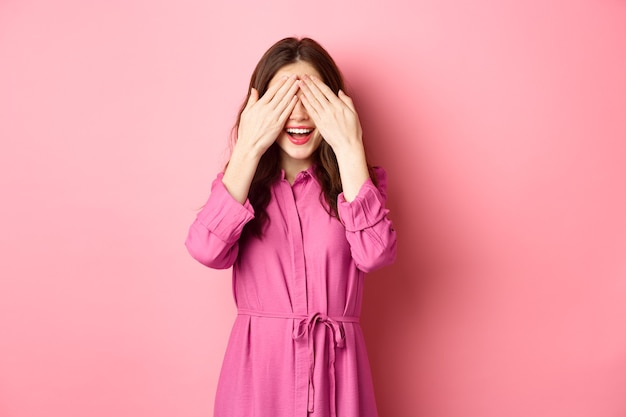 Enthousiaste femme brune en attente de cadeau surprise, couvrant les yeux et souriant, attendant présent, debout contre le mur rose.