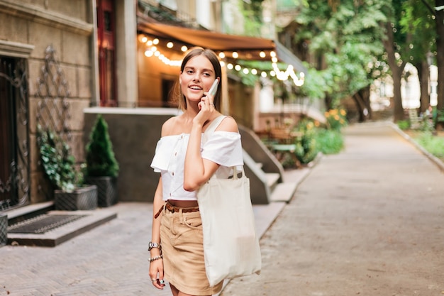 Enthousiaste femme charismatique parlant au téléphone tout en marchant le soir le long de la vieille rue