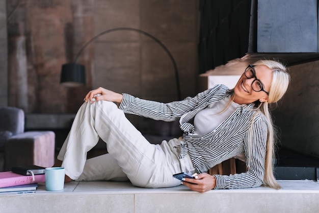 Enthousiaste femme designer dans des verres en riant à la maison tenant un téléphone souriant large Femme suédoise joyeuse en pantalon blanc et chemise heureuse de se détendre à la maison en s'amusant