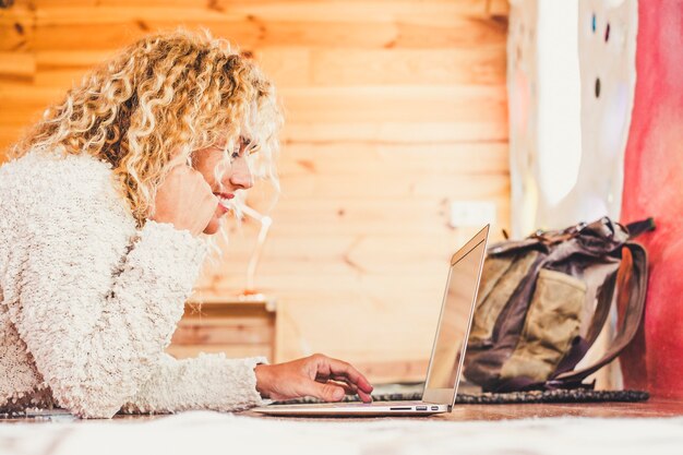 Enthousiaste heureuse belle blonde frisée caucasienne femme travaillant à l'ordinateur portable se coucha sur le sol à la maison ou à l'hôtel pour les vacances de voyage bureau alternatif pour un emploi gratuit et les voyages