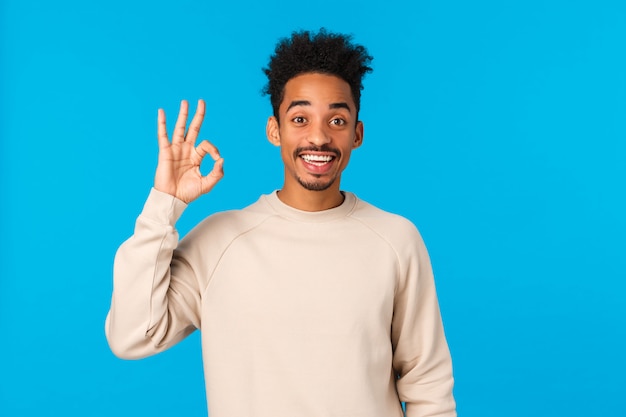 Enthousiaste heureux souriant afro-américain avec coupe de cheveux hipster, montrant un geste correct et signez la tête en accord, donnez votre approbation, confirmez ou acceptez de participer, debout mur bleu satisfait