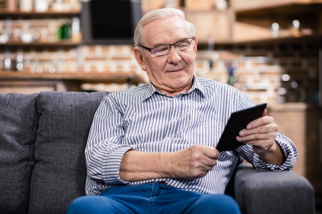 Enthousiaste homme âgé agréable assis sur le canapé et lisant un livre électronique
