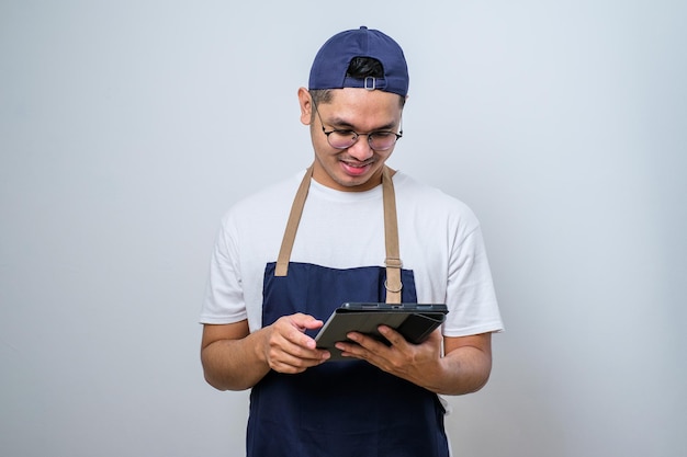 Enthousiaste jeune barista asiatique portant un tablier et une casquette à l'aide d'une tablette numérique pour gérer la commande de café