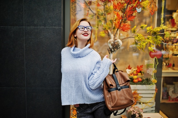 Enthousiaste jeune belle femme rousse dans des verres, pull en laine bleu chaud avec sac à dos posé en plein air contre les feuilles de l'automne, décorations d'arbres sur le magasin.