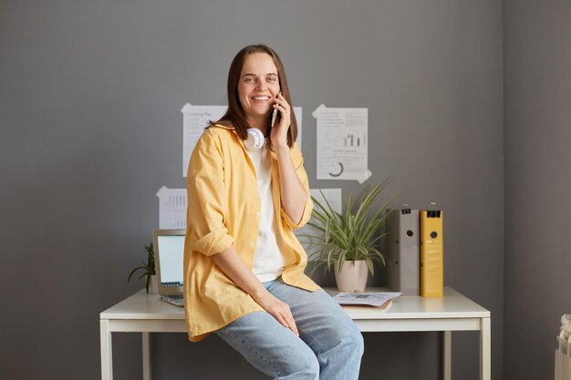 Enthousiaste jeune belle femme en veste jaune et jeans parlant au téléphone portable avec le sourire tout en étant assis sur une table au bureau ayant une pause en regardant la caméra avec un visage heureux