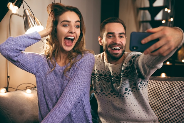 Enthousiaste jeune couple assis ensemble sur un canapé à la maison, prenant un selfie