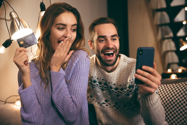 Enthousiaste jeune couple assis ensemble sur un canapé à la maison, tenant un téléphone mobile, montrant une carte de crédit en plastique