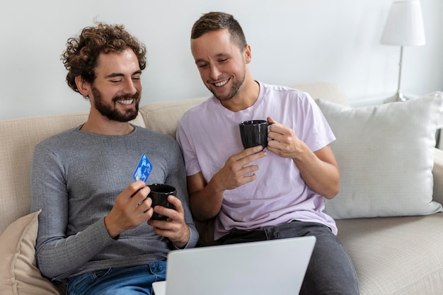 Enthousiaste jeune couple gay souriant joyeusement tout en faisant des achats en ligne à la maison Deux jeunes amants masculins utilisant une carte de crédit et un ordinateur portable pour effectuer un achat en ligne Jeune couple gay assis ensemble à l'intérieur