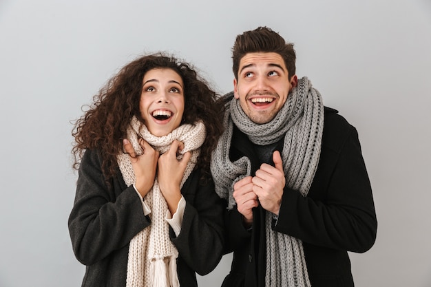 Enthousiaste jeune couple portant des chandails et des écharpes debout isolé sur mur gris, levant les yeux