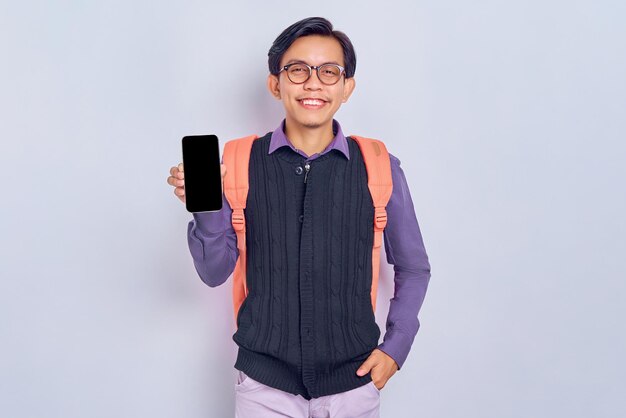 Enthousiaste jeune étudiant asiatique en vêtements décontractés avec sac à dos montrant un téléphone portable à écran blanc isolé sur fond gris Concept de collège universitaire de lycée