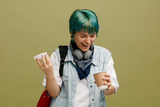 Enthousiaste jeune étudiante portant des écouteurs et un bandana sur le cou et le sac à dos tenant une tasse de café en papier et son capuchon regardant dans une tasse en riant isolé sur fond vert olive