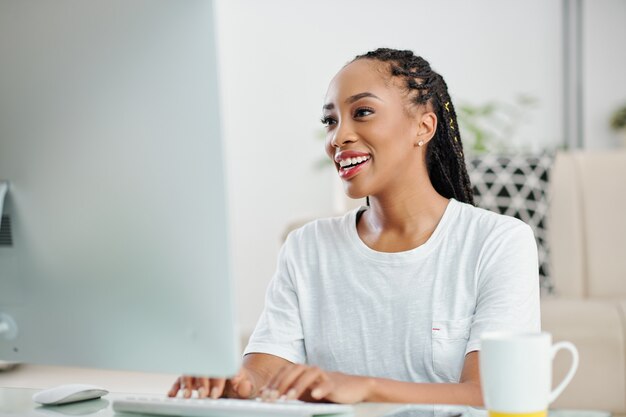 Enthousiaste jeune femme d'affaires noire assise au bureau et répondant aux e-mails de collègues et clients