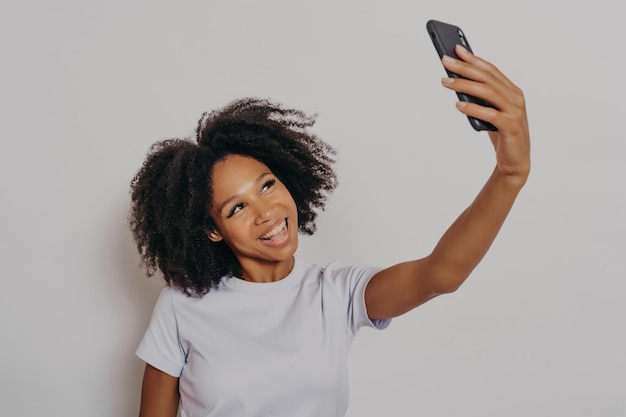 Enthousiaste jeune femme africaine aux cheveux bouclés prenant selfie sur son téléphone portable moderne