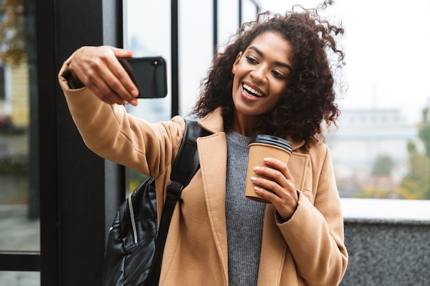 Enthousiaste jeune femme africaine portant manteau marchant à l'extérieur, tenant une tasse de café à emporter, prenant un selfie