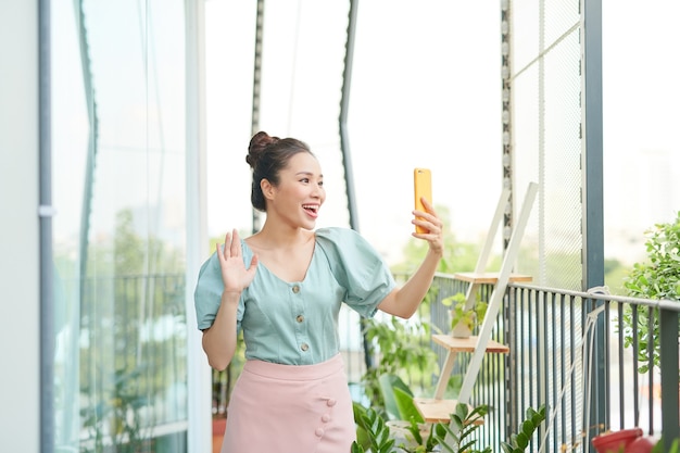 Enthousiaste jeune femme asiatique ayant un appel vidéo avec son téléphone en position debout sur un balcon