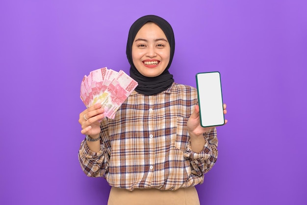 Enthousiaste jeune femme asiatique en chemise à carreaux montrant un téléphone portable à écran blanc et tenant des billets de banque isolés sur fond violet