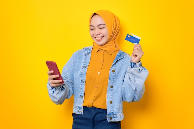 Enthousiaste jeune femme asiatique en veste de jeans tenant un téléphone portable et des achats en ligne par carte de crédit isolés sur fond jaune