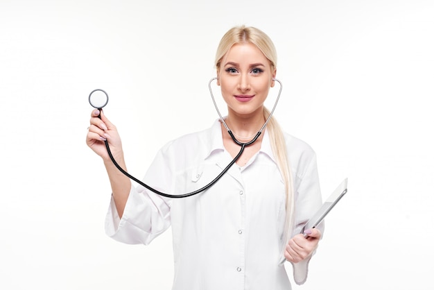 Enthousiaste jeune femme blonde médecin avec stéthoscope sur le cou en regardant la caméra isolée sur blanc. Tenant un stéthoscope à la main et écoutant le souffle