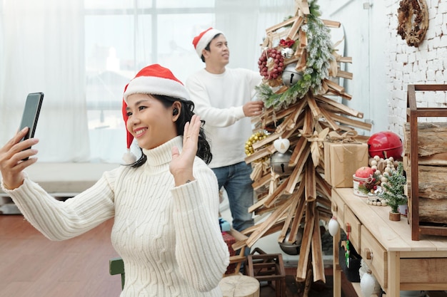 Enthousiaste jeune femme en bonnet de noel vidéo appelant des amis lorsque son mari décore le sapin de Noël en arrière-plan