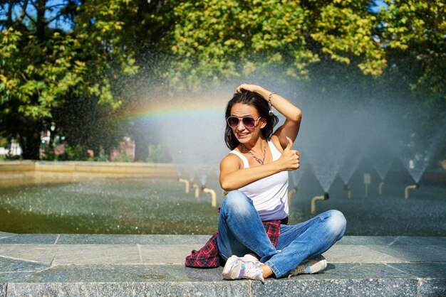 Enthousiaste jeune femme brune sur fond fontaine heureux montre le geste