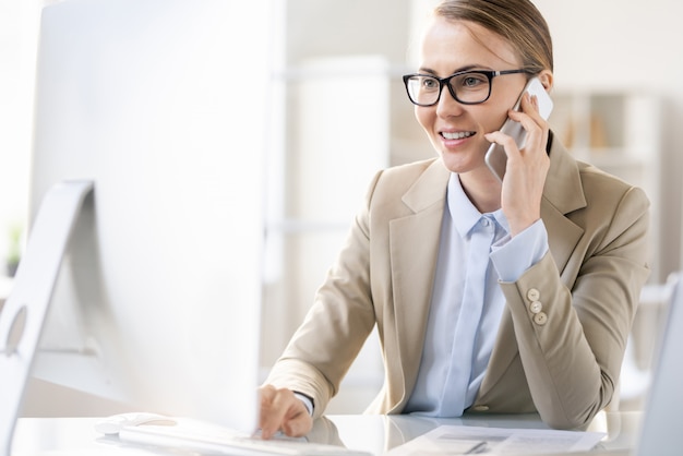 Enthousiaste jeune femme confiante dans des verres assis à table et répondre à un appel tout en utilisant un ordinateur dans un bureau moderne