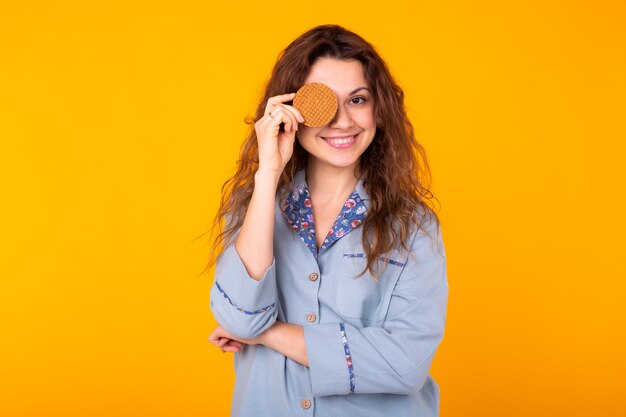 Enthousiaste jeune femme couvrant son œil avec cookie. Elle rit de joie. Mur jaune.