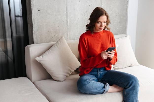 Enthousiaste jeune femme habillée avec désinvolture assis sur un canapé à la maison, tenant un téléphone mobile