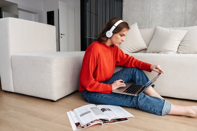 Enthousiaste jeune femme habillée avec désinvolture dans des écouteurs assis sur un sol à la maison, étudiant avec un ordinateur portable