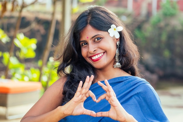 Enthousiaste jeune femme indienne avec du flover blanc dans les cheveux montrant la forme de coeur avec les mains relaxantes sur la plage à Goa