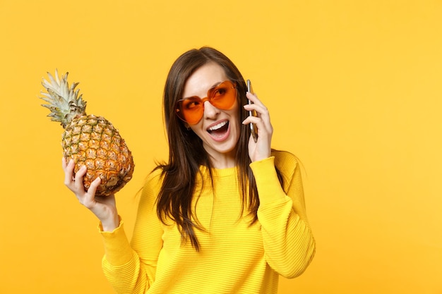 Enthousiaste jeune femme en lunettes de soleil coeur tenant des fruits mûrs d'ananas, parlant au téléphone mobile isolé sur fond orange jaune. Mode de vie vivant des gens, concept de vacances relaxantes. Espace de copie maquette