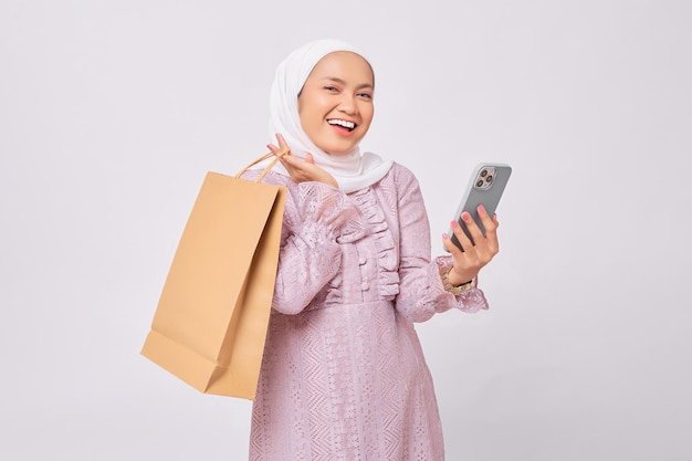 Enthousiaste jeune femme musulmane asiatique portant le hijab et la robe violette tenant un sac à provisions et un téléphone portable isolés sur fond de studio blanc