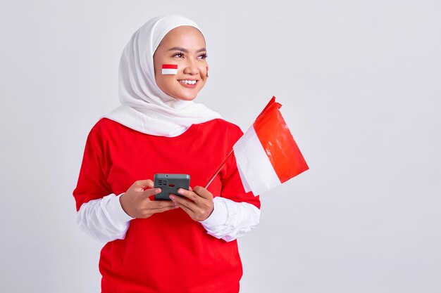Enthousiaste jeune femme musulmane asiatique en tshirt blanc rouge à l'aide de téléphone portable et célébrant le jour de l'indépendance indonésienne le 17 août isolé sur fond blanc