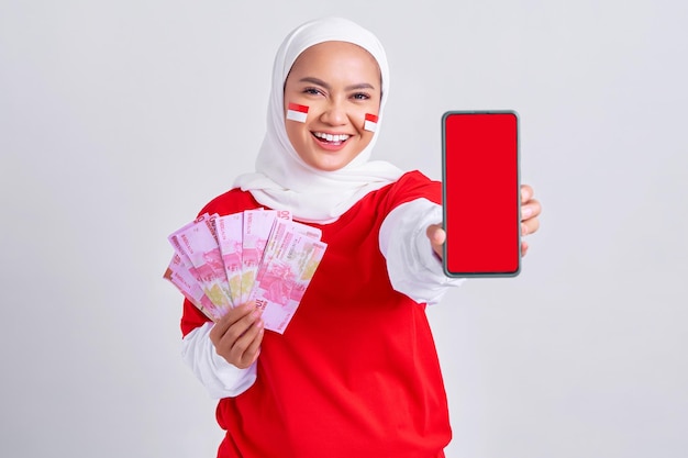 Enthousiaste jeune femme musulmane asiatique en tshirt blanc rouge montrant un téléphone portable et de l'argent en espèces en billets de roupie indonésienne isolés sur fond blanc Jour de l'indépendance indonésienne le 17 août concept