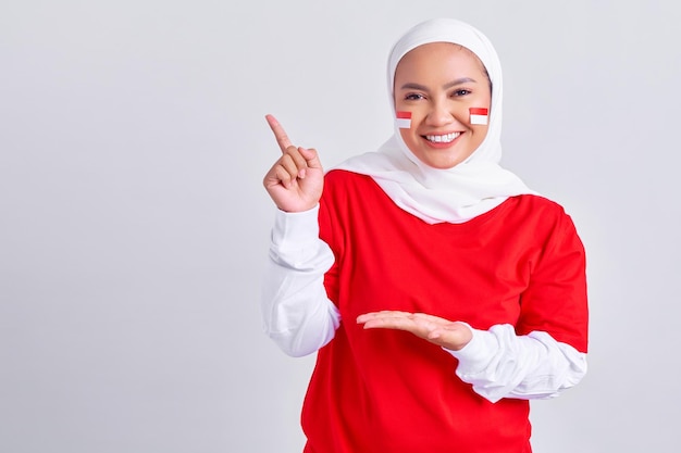 Enthousiaste jeune femme musulmane asiatique en tshirt blanc rouge pointant de côté à l'espace de copie avec le doigt et la paume isolé sur fond blanc Fête de l'indépendance indonésienne le 17 août concept