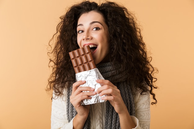 Enthousiaste jeune femme portant chandail et écharpe tenant une grande barre de chocolat isolée sur mur beige