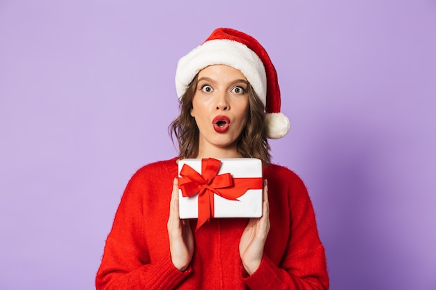 Enthousiaste jeune femme portant un chapeau de Noël rouge debout isolé sur mur violet, tenant une boîte-cadeau