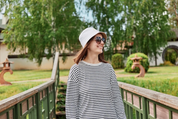 Enthousiaste jeune femme portant une chemise rayée et un panama se reposant sur un parc debout sur un pont profitant du temps d'été fille souriante à lunettes de soleil