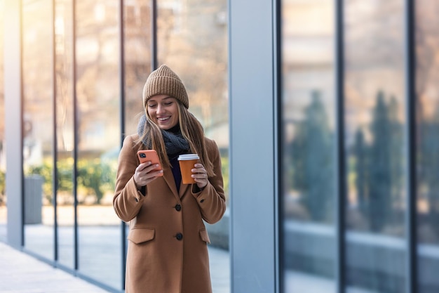 Enthousiaste jeune femme portant un manteau marchant à l'extérieur tenant une tasse de café à emporter à l'aide d'un téléphone portable