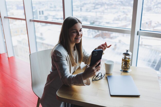 Enthousiaste jeune femme riant en lisant une notification amusante sur un téléphone intelligent assis devant un ordinateur portable dans un café