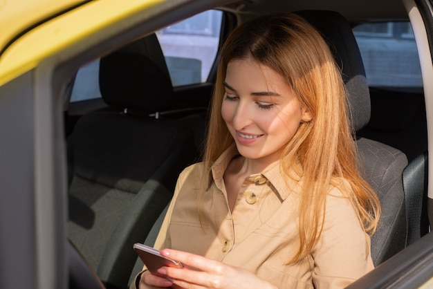 Photo enthousiaste jeune femme vérifiant son smartphone alors qu'il était assis dans le siège du conducteur de sa voiture jaune