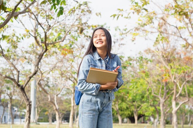 Enthousiaste jeune fille à l'aide d'un téléphone portable et tenant un livre debout dans le parc porte un concept de mode de vie veste et jeans