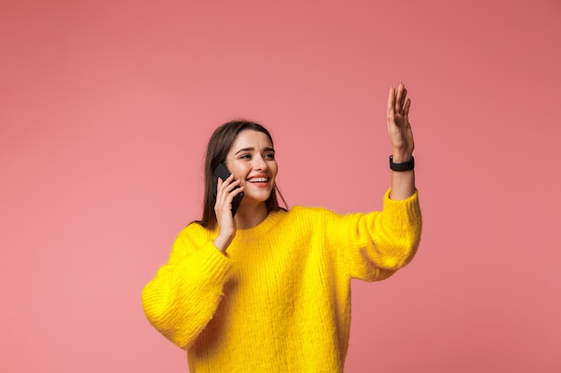 Enthousiaste jeune fille portant un chandail debout isolé sur rose, parler au téléphone mobile, en agitant