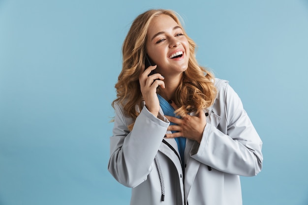 Enthousiaste jeune fille portant un imperméable debout isolé, parler au téléphone mobile