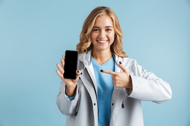 Enthousiaste jeune fille portant un imperméable debout isolé, pointant le doigt sur un téléphone mobile à écran blanc