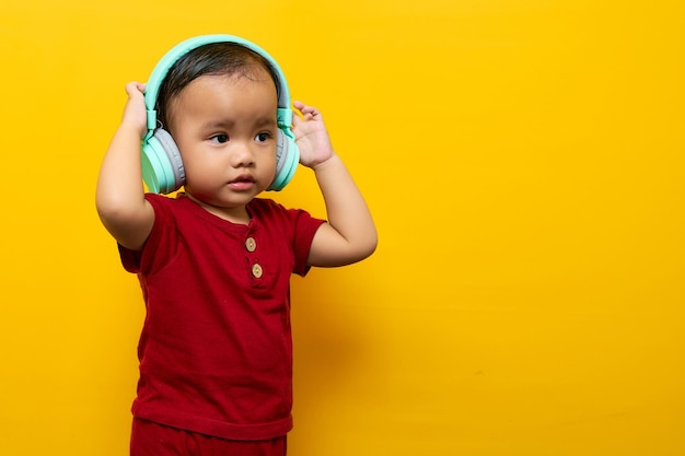 Enthousiaste jeune garçon asiatique 2 ans bébé garçon en t-shirt rouge Écouter de la musique préférée avec un casque isolé sur fond jaune Concept de mode de vie familial amour fête des mères