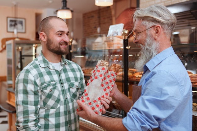 Enthousiaste jeune homme achetant du pain frais d'un boulanger senior au magasin de pain local