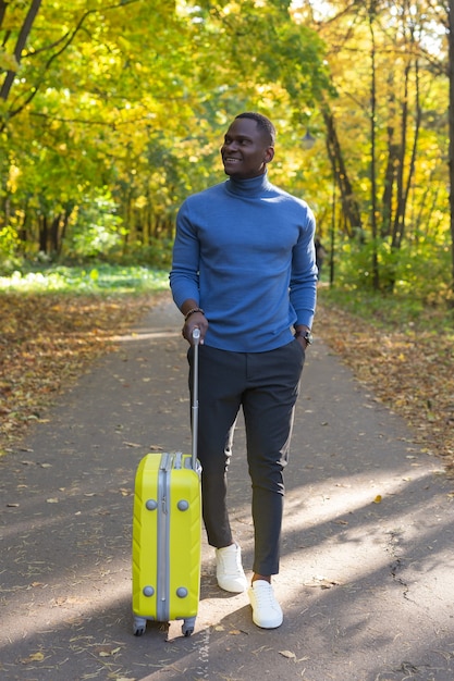 Enthousiaste jeune homme afro-américain dans des vêtements élégants avec valise se promène dans un parc d'automne par une journée chaude.