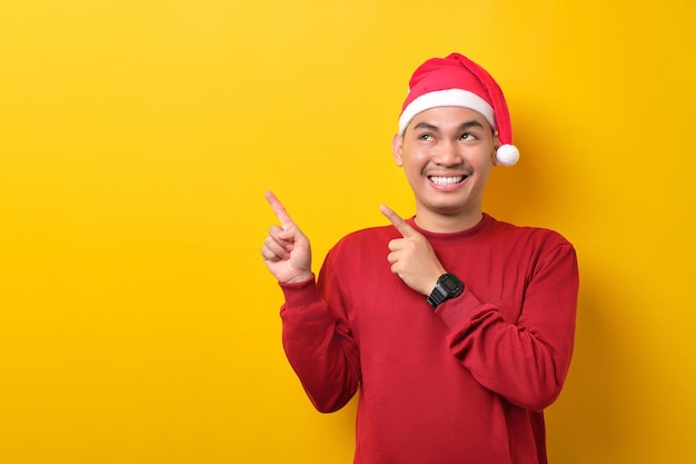 Enthousiaste jeune homme asiatique en bonnet de noel pointant les doigts de côté sur un espace vide pour le texte publicitaire sur fond de studio jaune célébration vacances de Noël et concept du nouvel an