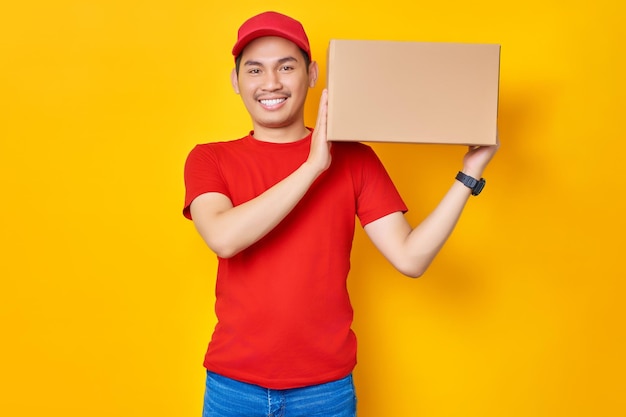 Enthousiaste jeune homme asiatique en bonnet rouge tshirt uniforme employé travaille comme concessionnaire courrier transportant la boîte de colis à l'épaule isolé sur fond jaune Concept de service de livraison professionnel